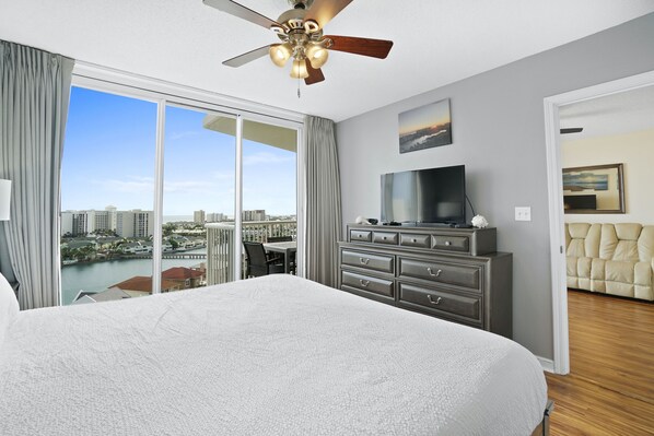 A hotel room with a large bed, a ceiling fan, a TV, a dresser, and a city view through the window.