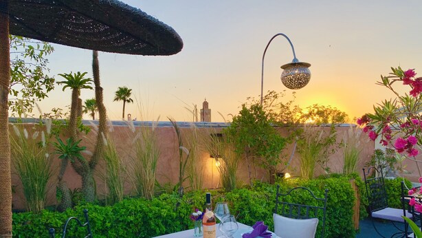 An outdoor dining area with a table set for two, surrounded by lush greenery and a view of a cityscape at sunset. - Riad 11 Zitoune (Marrakech)