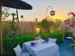 An outdoor dining area with a table set for two, surrounded by lush greenery and a view of a cityscape at sunset. - Riad 11 Zitoune (Marrakech)