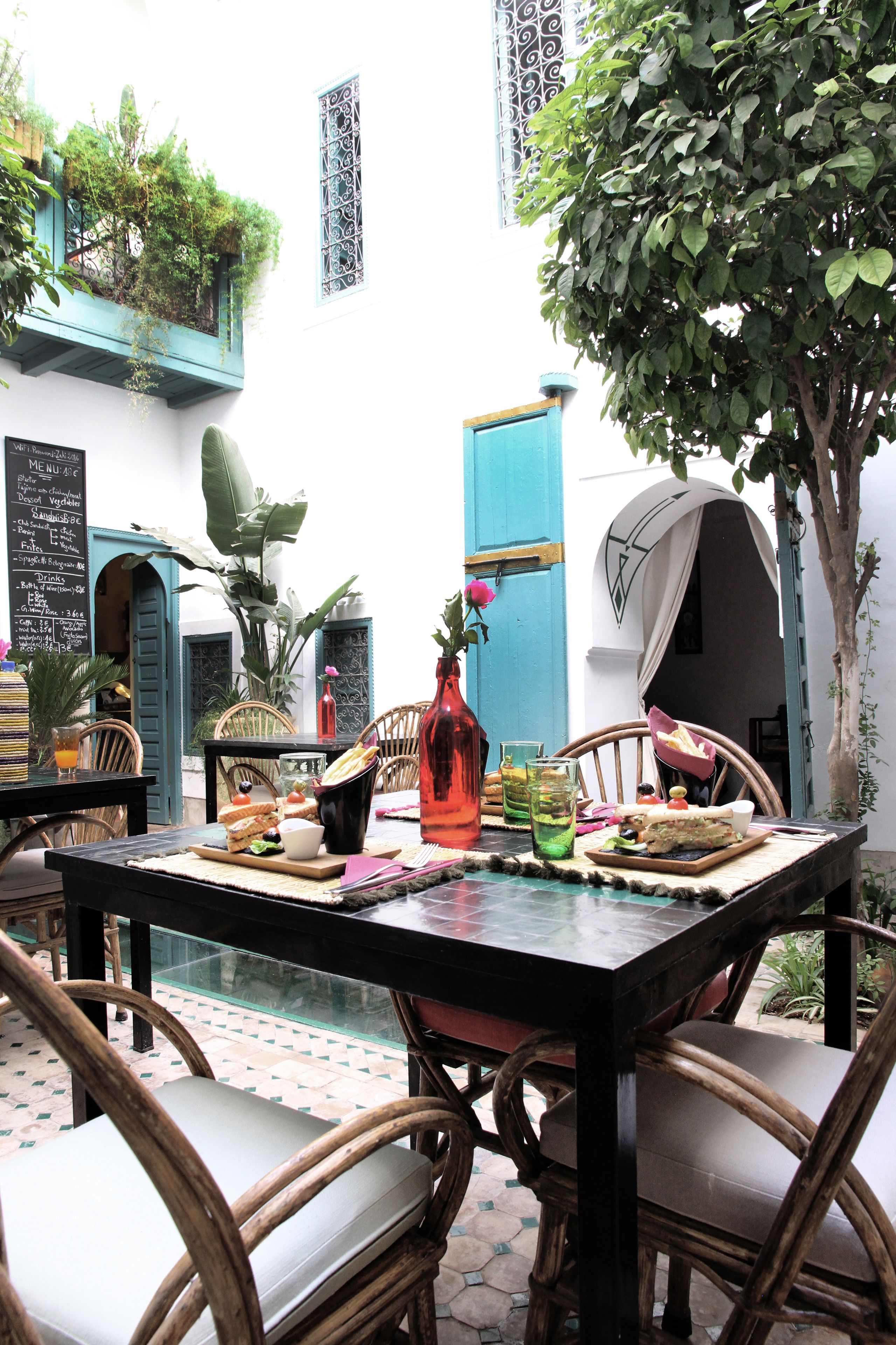 An outdoor café with a table set for two, surrounded by wicker chairs, a menu board, and a potted plant.