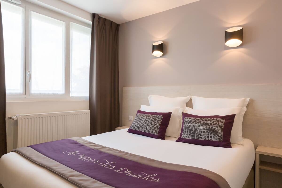 Una cama bien hecha con una colcha morada y dos almohadas estampadas, una ventana con cortinas y dos lámparas de pared.