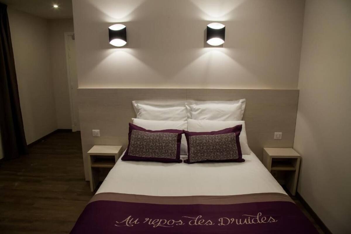 Una cama bien tendida con dos almohadas, dos mesitas de noche y dos lámparas de pared.