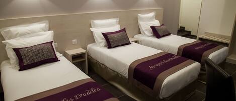 Habitación de hotel con tres camas, cada una con cabecero blanco y manta morada. Hay luces fijadas a la pared sobre las camas y un espejo en la pared.