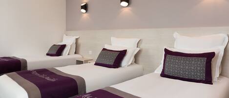 Habitación de hotel con tres camas, cada una con una manta morada y un almohadón. Hay luces fijadas a la pared sobre las camas.