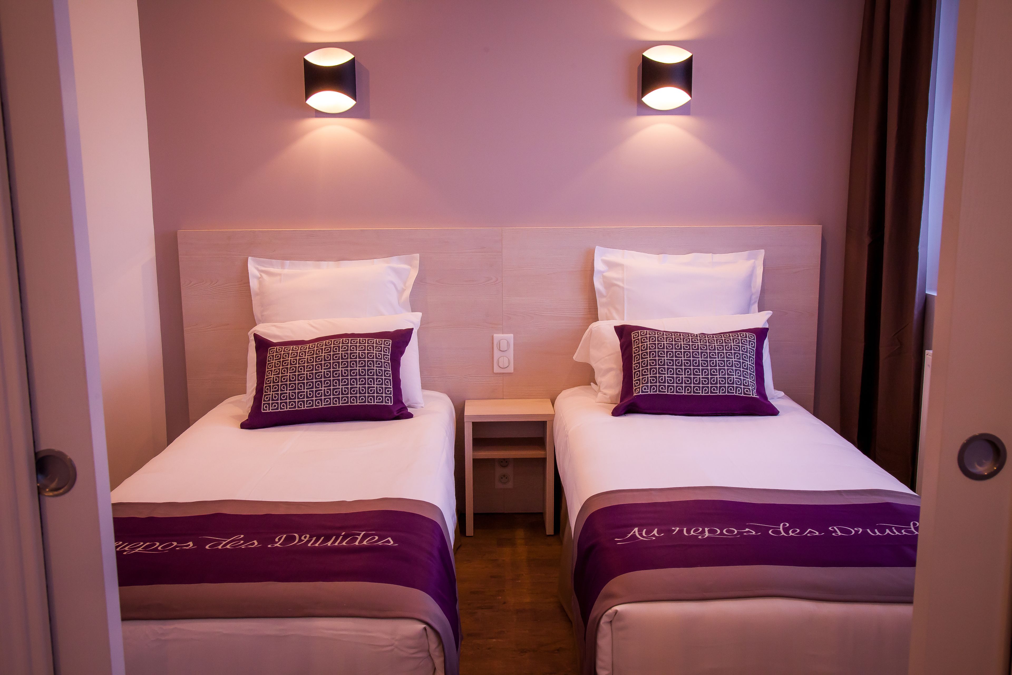 A hotel room with two single beds, each with a headboard, a nightstand, and a wall-mounted light fixture.