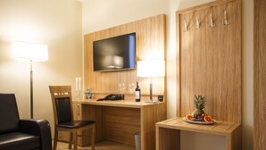 Quarto de hotel com uma escrivaninha de madeira, uma cadeira, uma TV fixada na parede, uma pequena mesa com fruta e uma luminária.