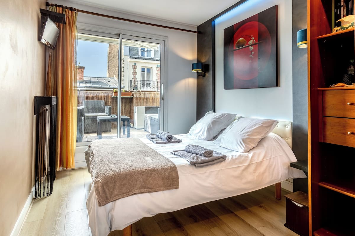 A hotel room with a bed, a wooden floor, a sliding glass door to an outdoor balcony, a wall-mounted TV, and a bookshelf.