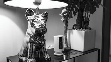 Foto em preto e branco de uma estatueta de cachorro decorativa sobre uma mesa, uma luminária, um pequeno aparelho branco e um vaso com planta.