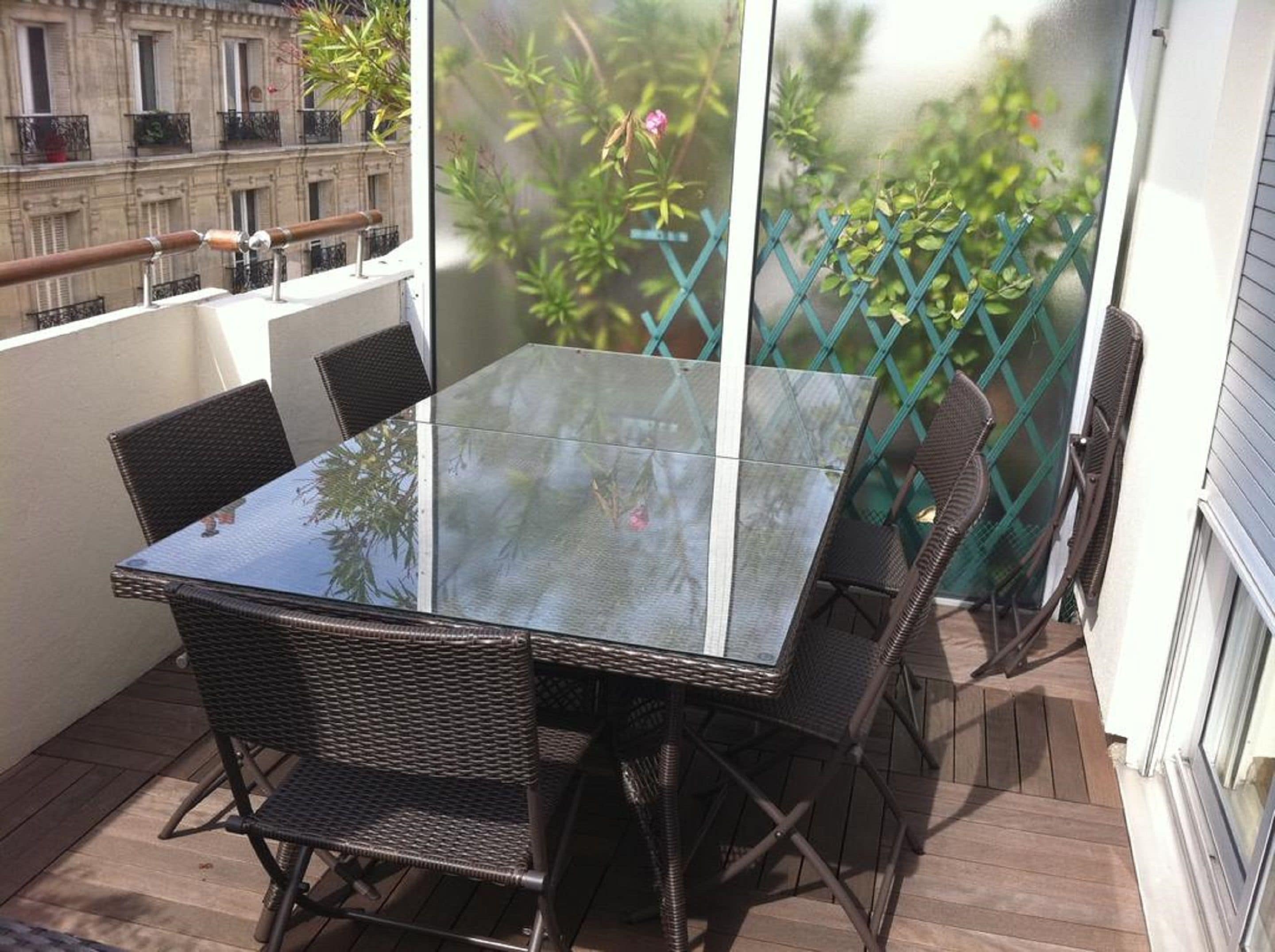 Una mesa con cubierta de vidrio y sillas de mimbre en un balcón con vista a follaje y un edificio al fondo.