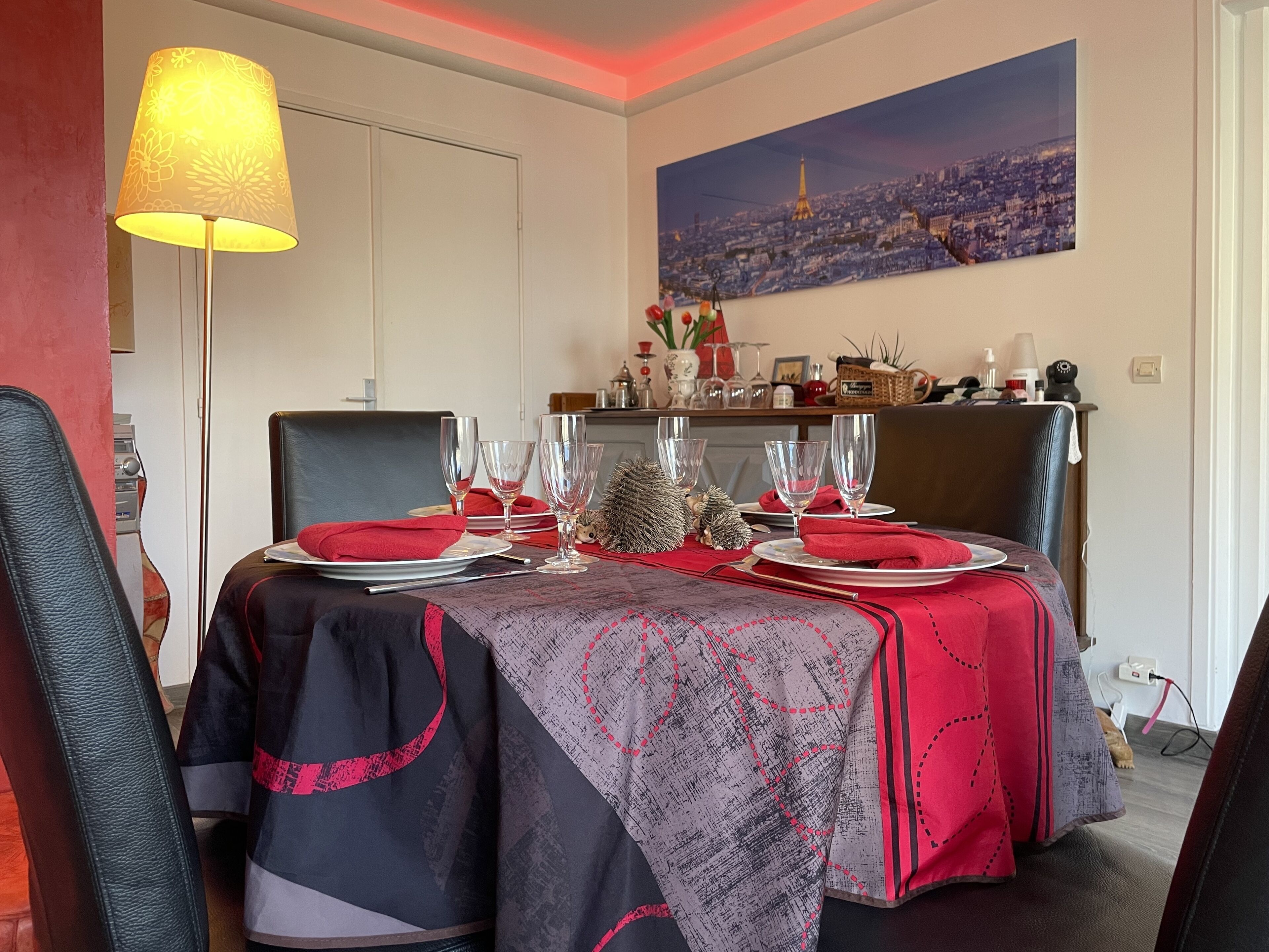 Un coin repas avec deux tables dressées pour le repas, une lampe et un tableau représentant un paysage urbain accroché au mur.