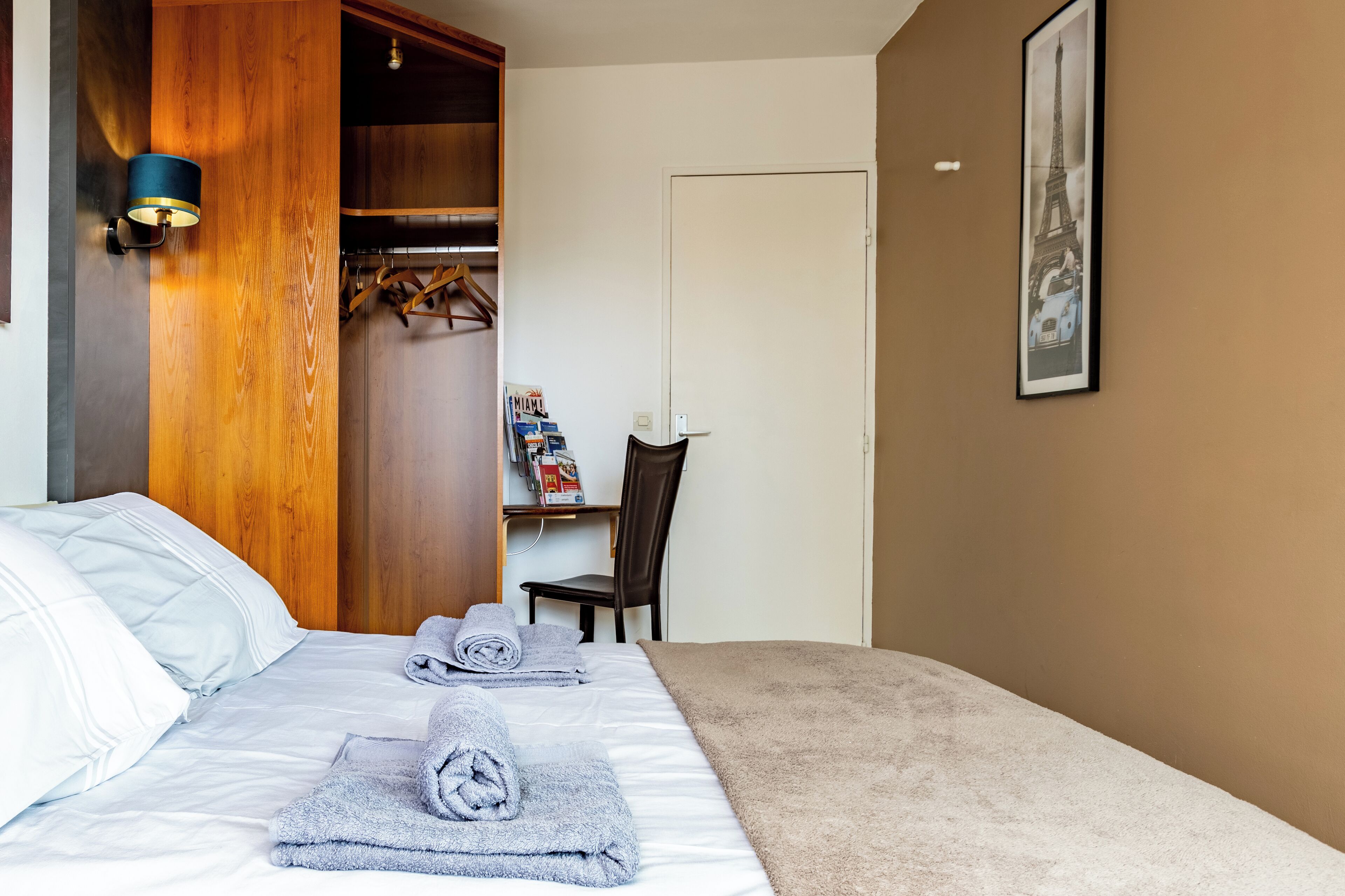 Una cama bien hecha con toallas, un armario de madera, un escritorio con silla y un cuadro en la pared.