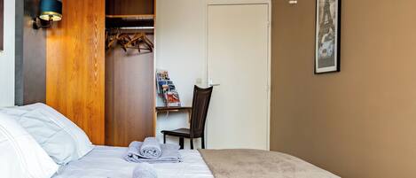 A neatly made bed with towels, a wooden wardrobe, a desk with a chair, and a framed picture on the wall.