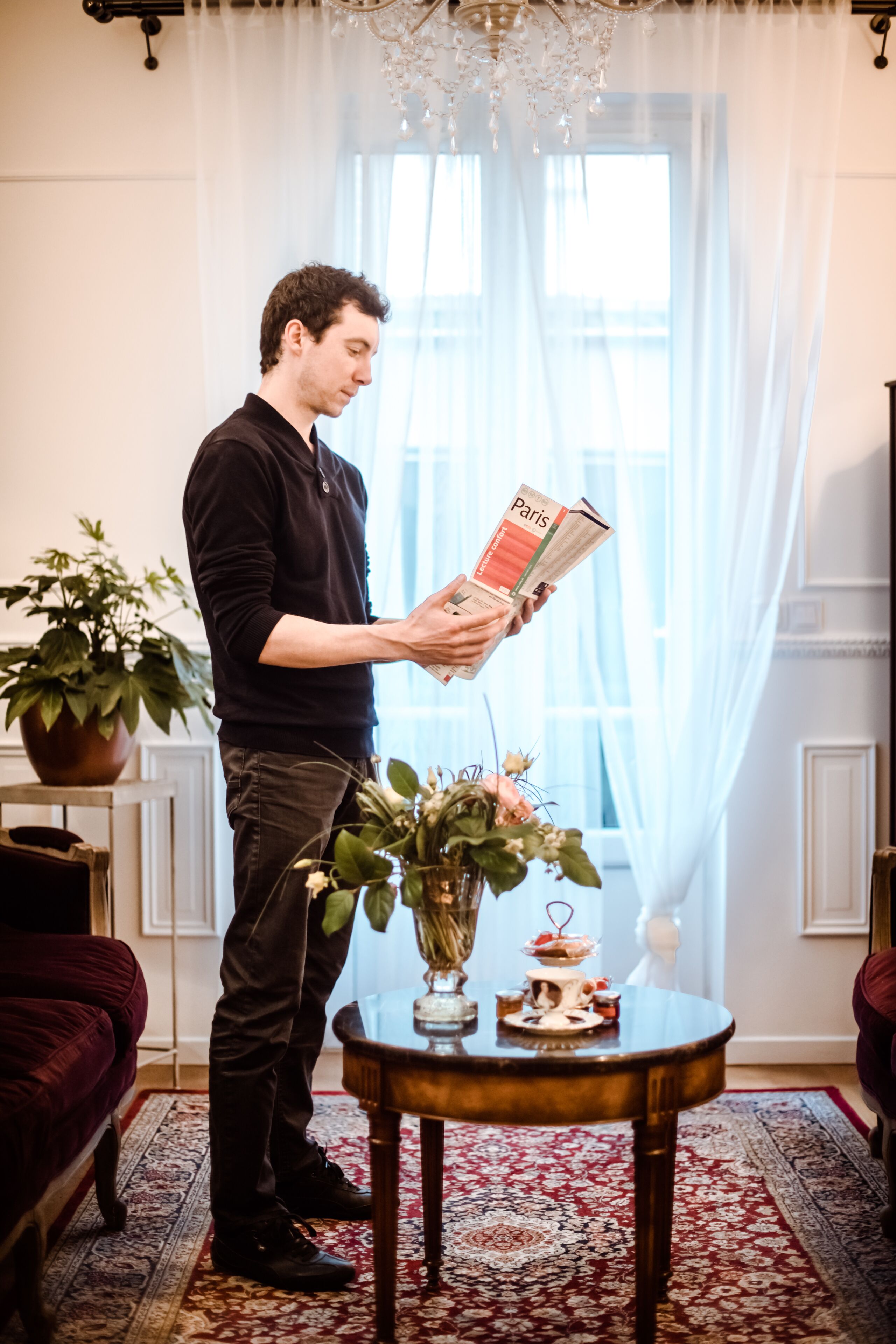 A man reading a magazine in a room with a large window, a chandelier, and a table with a vase of flowers.