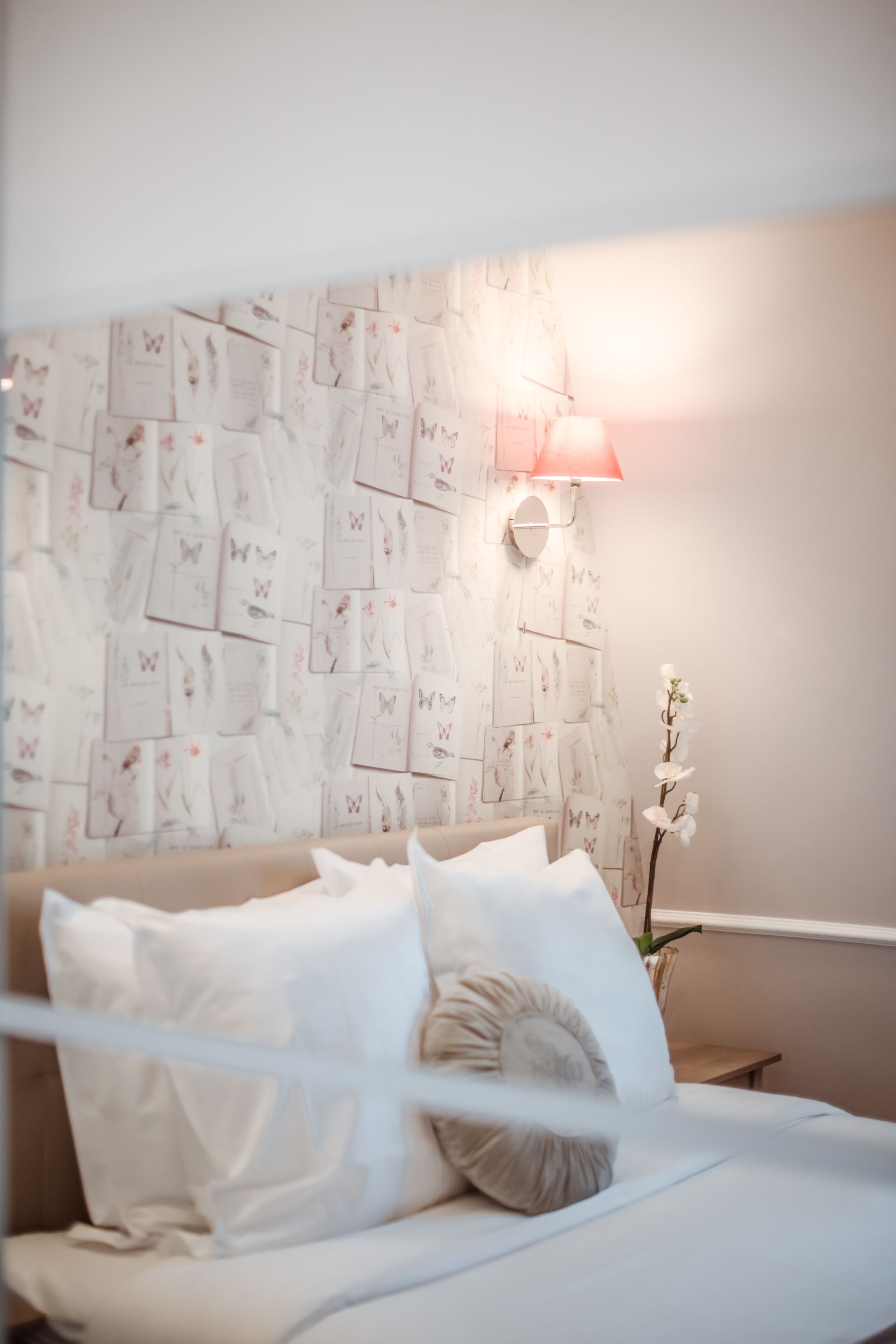 A bedroom with a bed, pillows, a wall with butterfly illustrations, a nightstand, and a lamp.