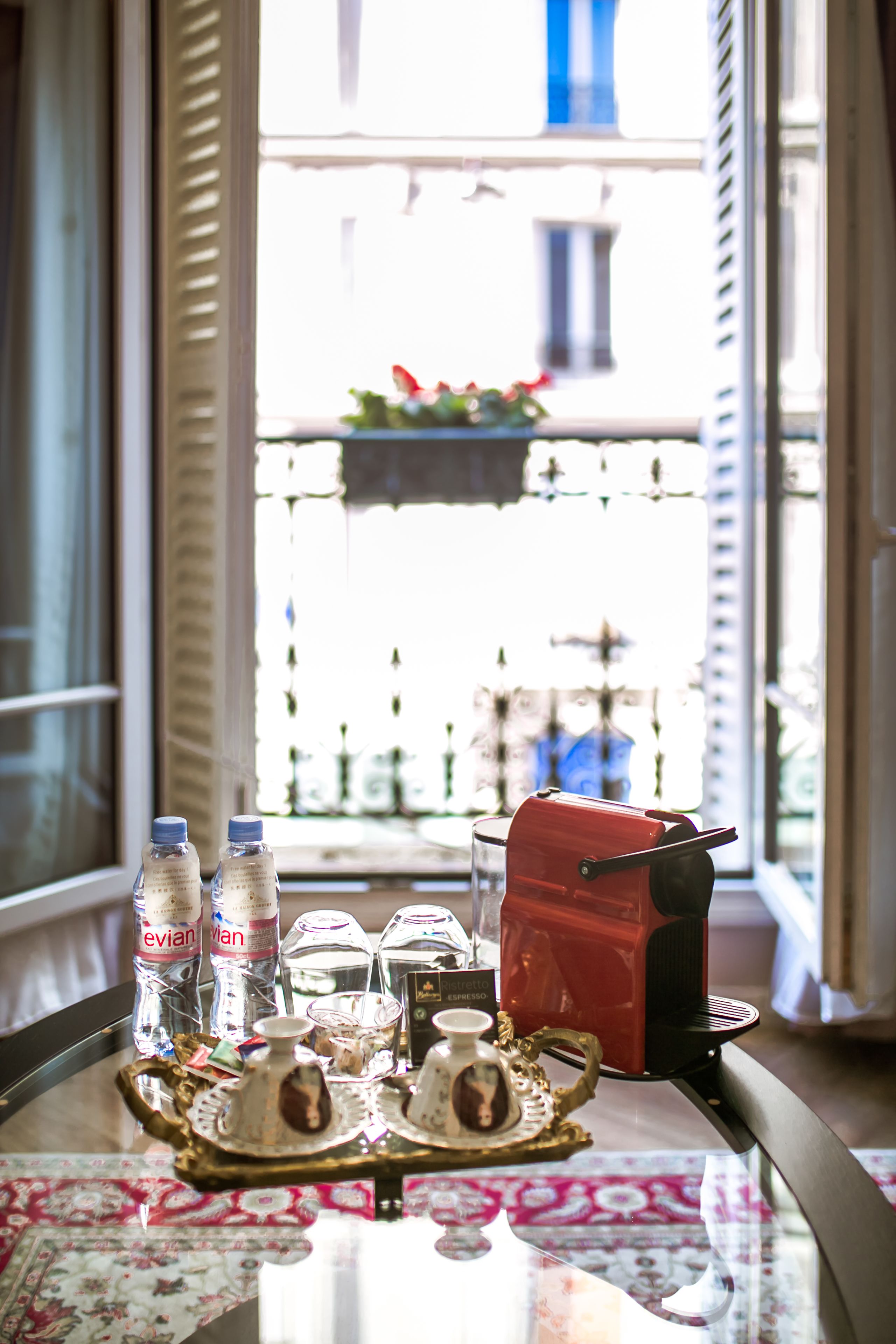Une table avec un plateau de boissons, une théière et un sac rouge, placée près d’une fenêtre donnant sur un balcon.