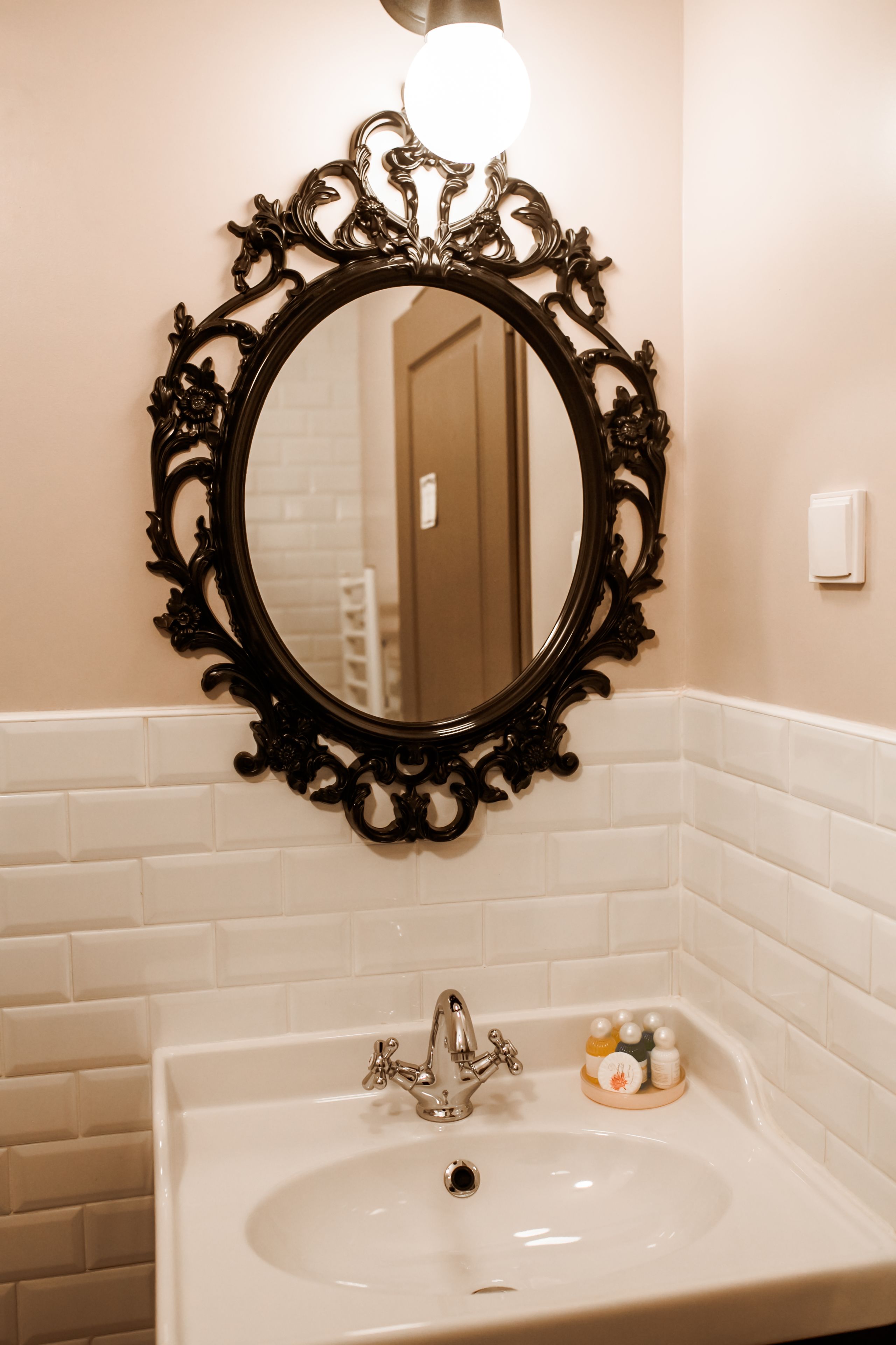 A bathroom with a white sink, ornate black mirror, and white tiled walls.
