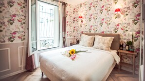 A bedroom with floral wallpaper, a bed with a white duvet, a nightstand with a lamp, and a window with curtains. - La Maison Gobert Paris Hotel Particulier (Paris)