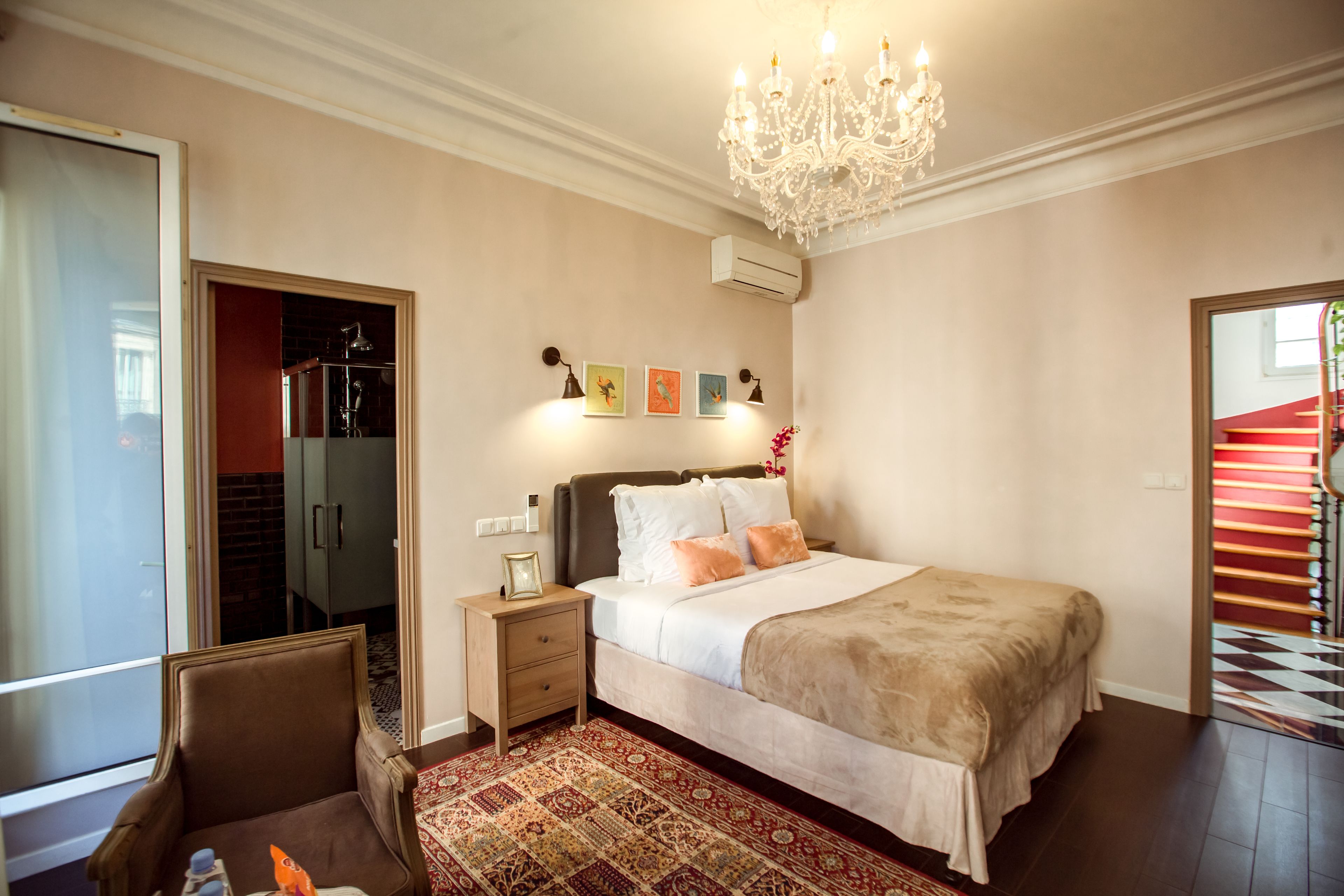 Une chambre à coucher avec un grand lit, un lustre, un tapis à motifs rouges et beiges, et un escalier en arrière-plan.