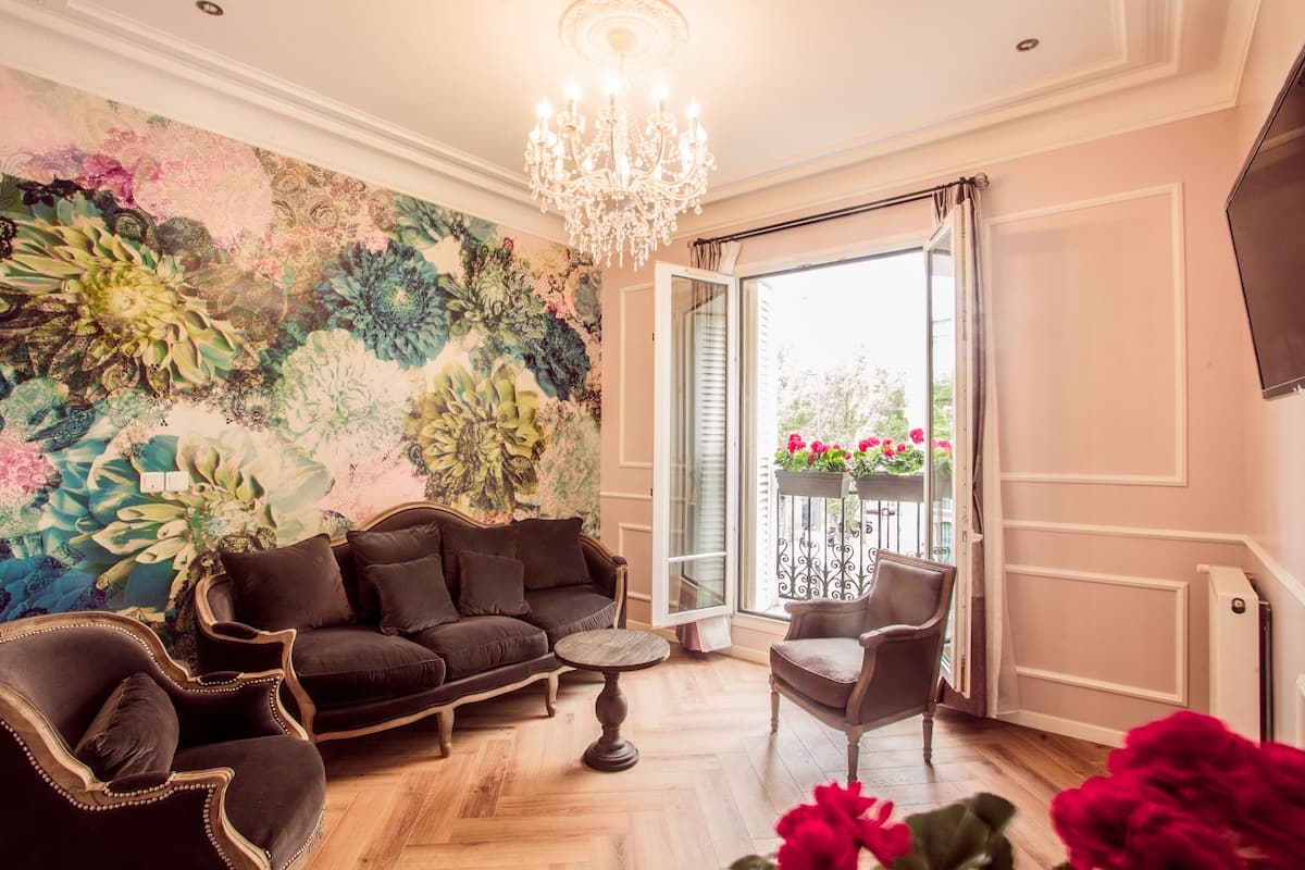A living room with a dark brown sofa, a round coffee table, and a chair. A large floral tapestry adorns the wall, and a chandelier hangs from the ceiling.