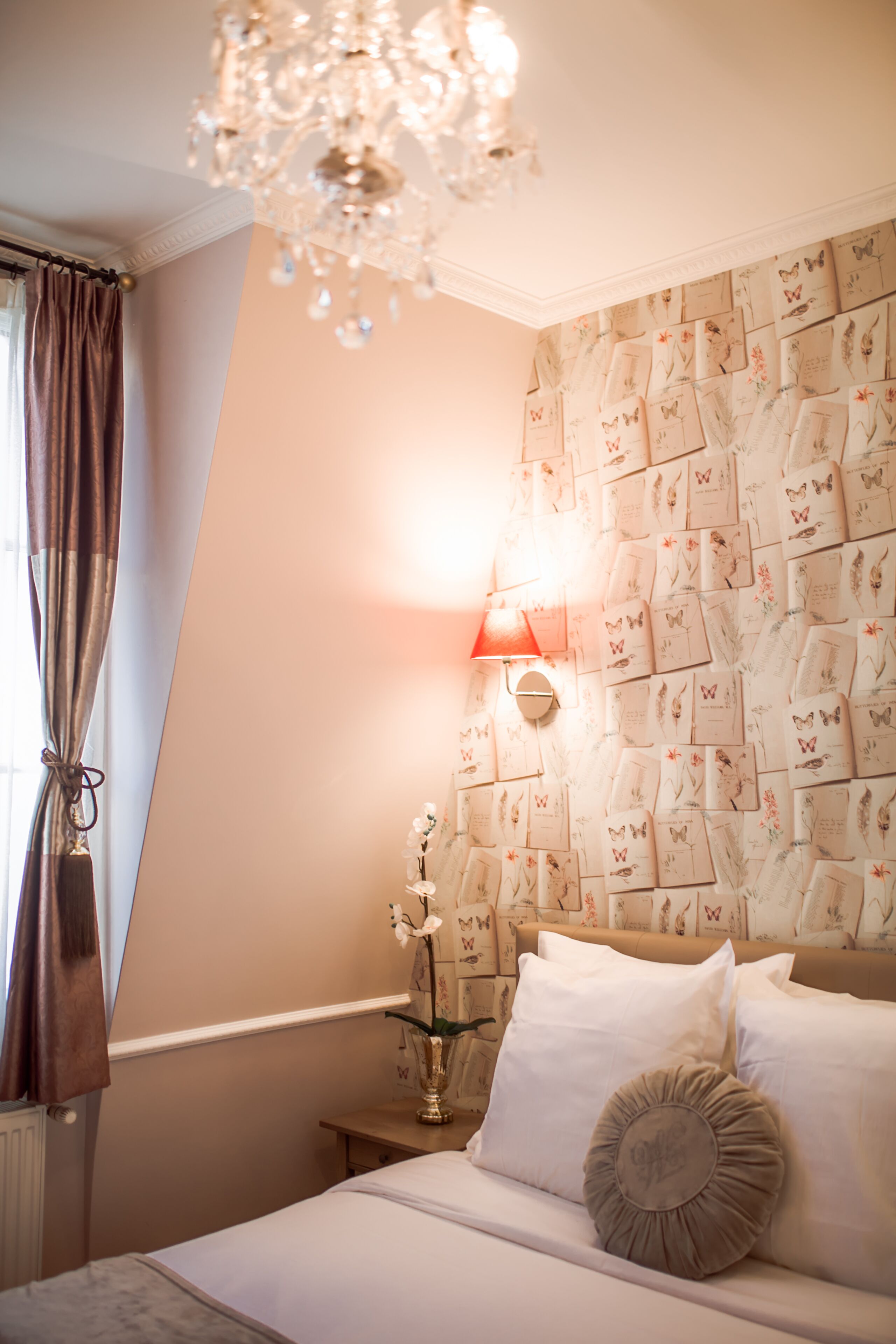 A bedroom with a bed, a chandelier, a wall-mounted lamp, and a floral wallpaper pattern.