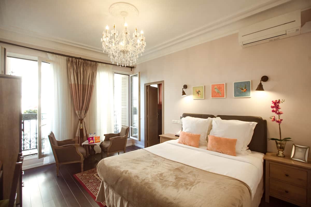 A hotel room with a large bed, a chandelier, a desk with a chair, a small table, and artwork on the wall.