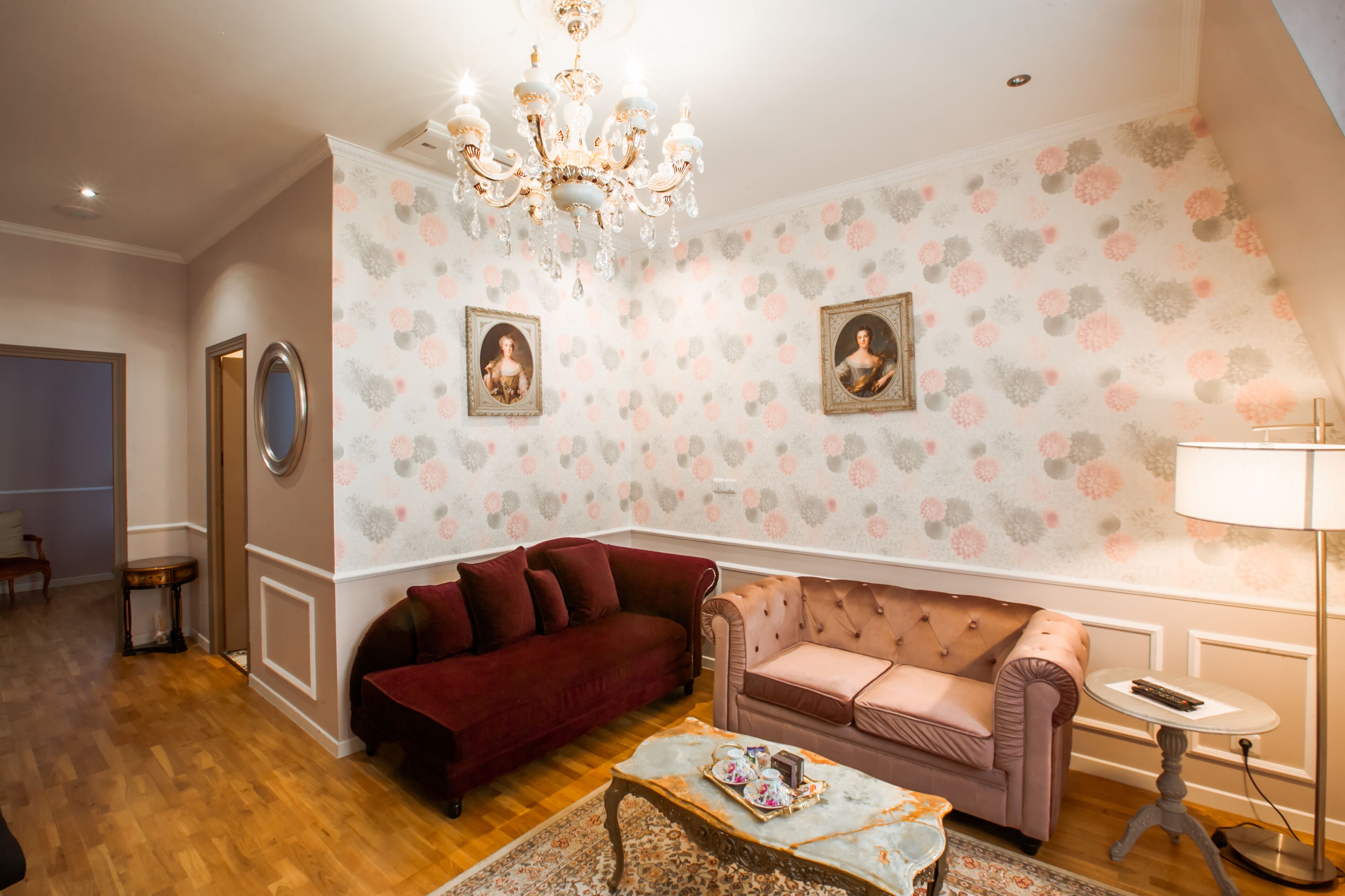 Een woonkamer met een rode bank, een roze bank en een salontafel. Twee ingelijste foto’s hangen aan de muur en er is een kroonluchter.