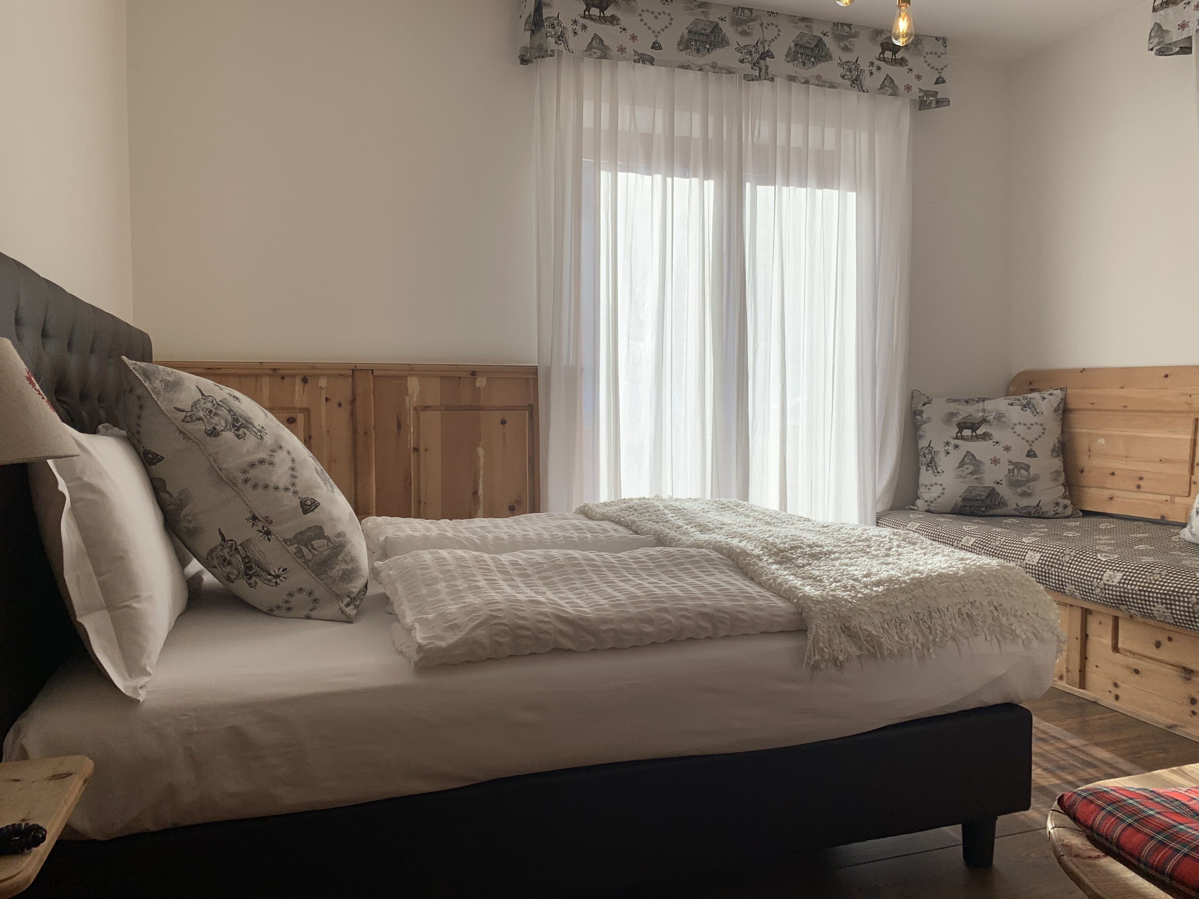 Ein Schlafzimmer mit einem Bett, einem hölzernen Kopfteil, einem Fenster mit durchsichtigen Vorhängen und einem Nachttisch.