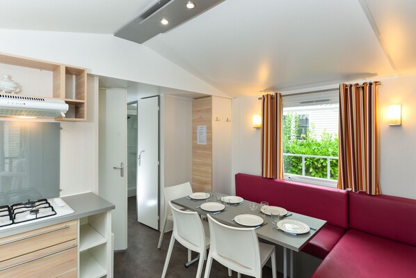 A compact kitchenette with a gas stove, a small refrigerator, and a built-in oven. There is a dining area with a table set for two, white chairs, and a red cushioned bench. A window with striped curtains allows natural light to enter the room. - Camping le Kergariou (Clohars-Carnoët)