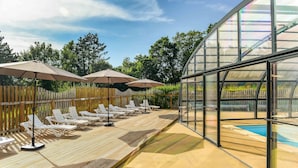 Un área junto a la piscina con tumbonas y sombrillas, una terraza de madera y una piscina cubierta con cristal.