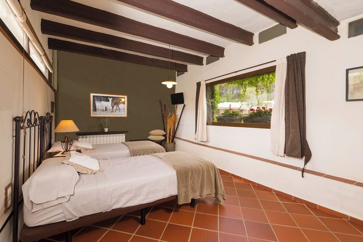 A room with two beds, a window with curtains, a picture on the wall, and a lamp.