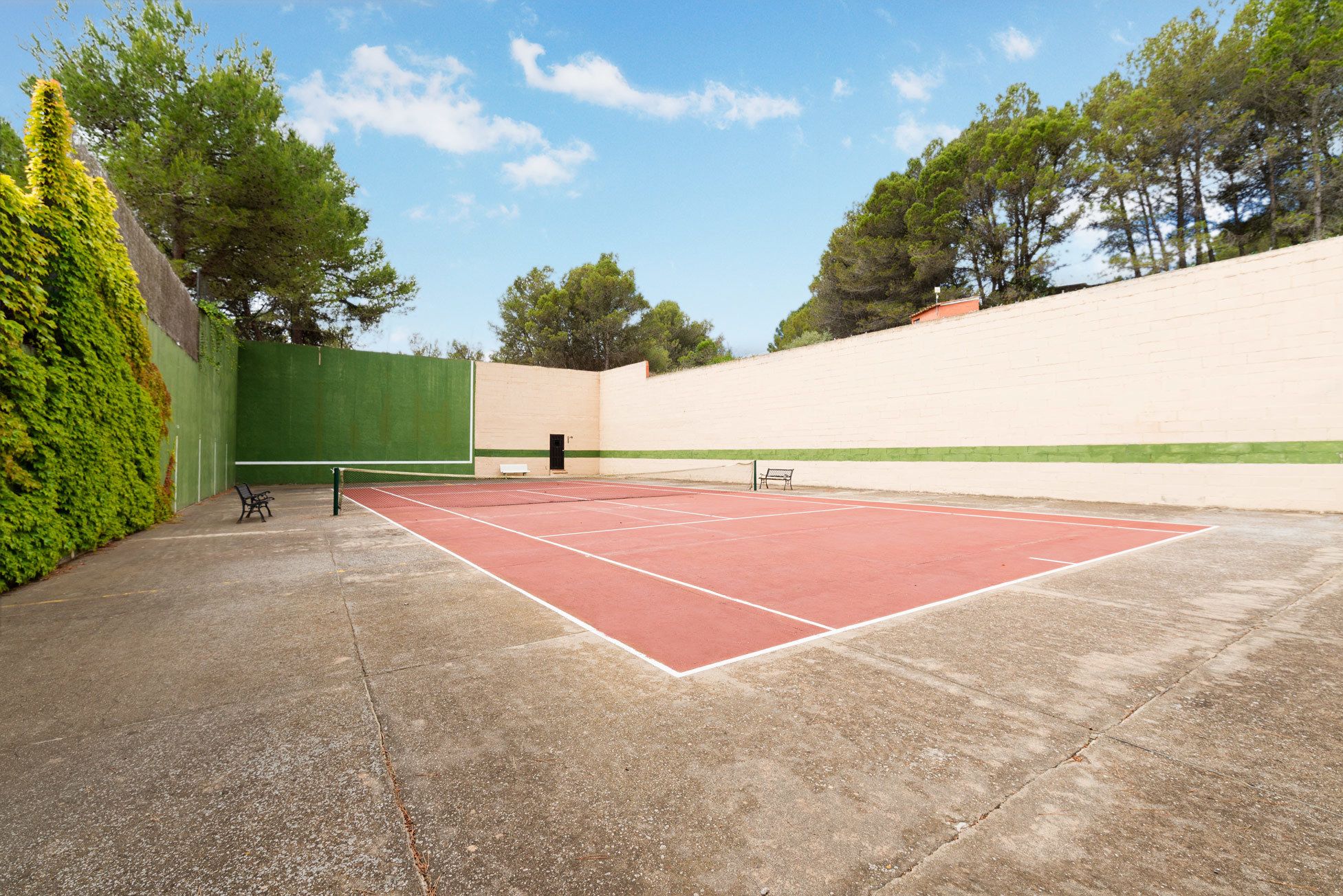 Una cancha de tenis al aire libre con una valla verde, un banco y árboles de fondo.