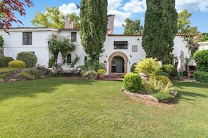 A white building with a stone archway entrance, surrounded by a well-maintained garden with various plants and a lawn. - Comarquinal Bioresort Penedès (Sant Quinti de Mediona)