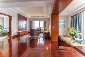 A spacious hotel room with a wooden floor, a large flat-screen TV, a comfortable seating area with a sofa and armchairs, a dining table with a vase of flowers, and a large window with curtains. - Glory Charm Hotel (Fangchenggang)