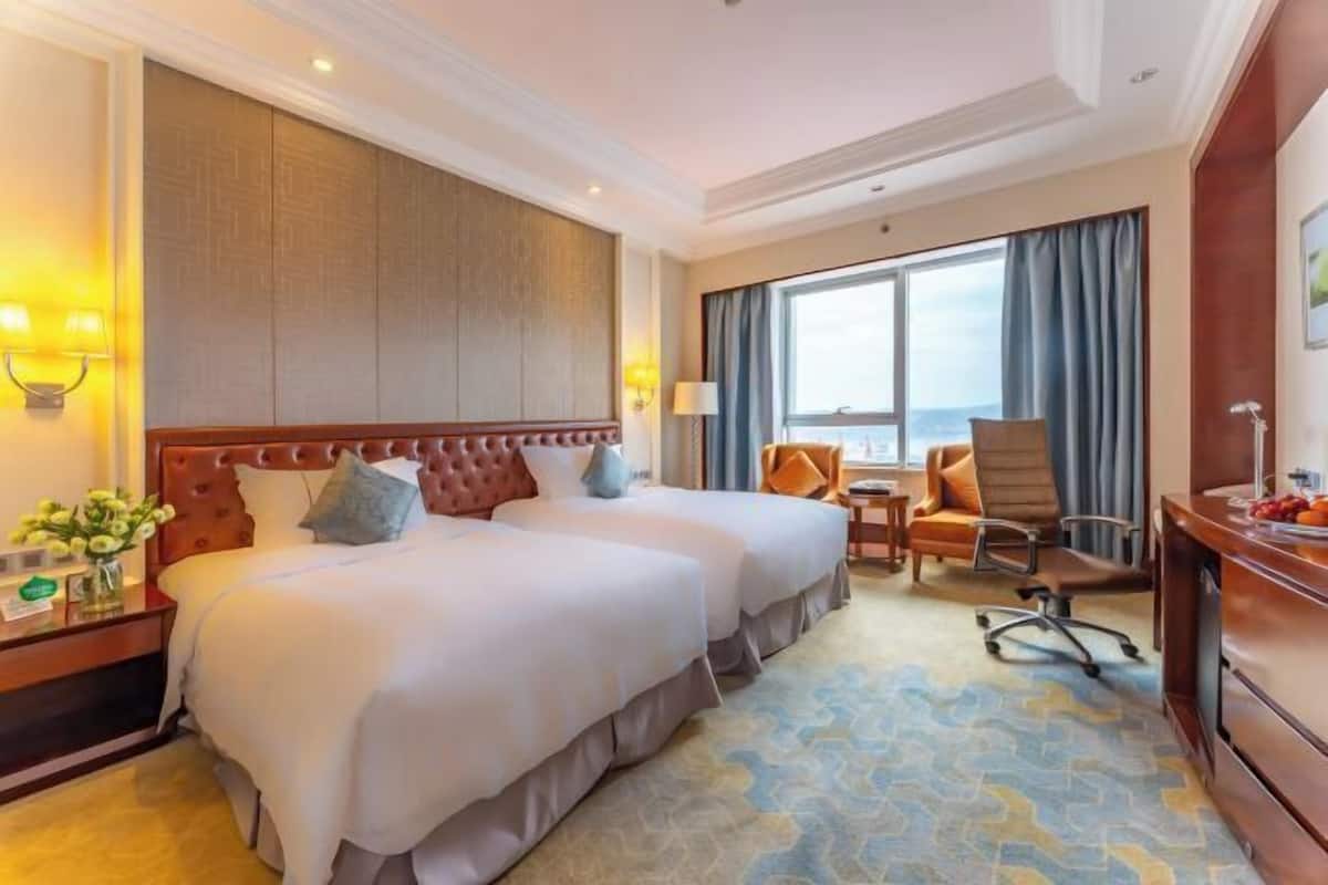 Quarto de hotel com duas camas, uma escrivaninha, uma cadeira e uma janela grande.