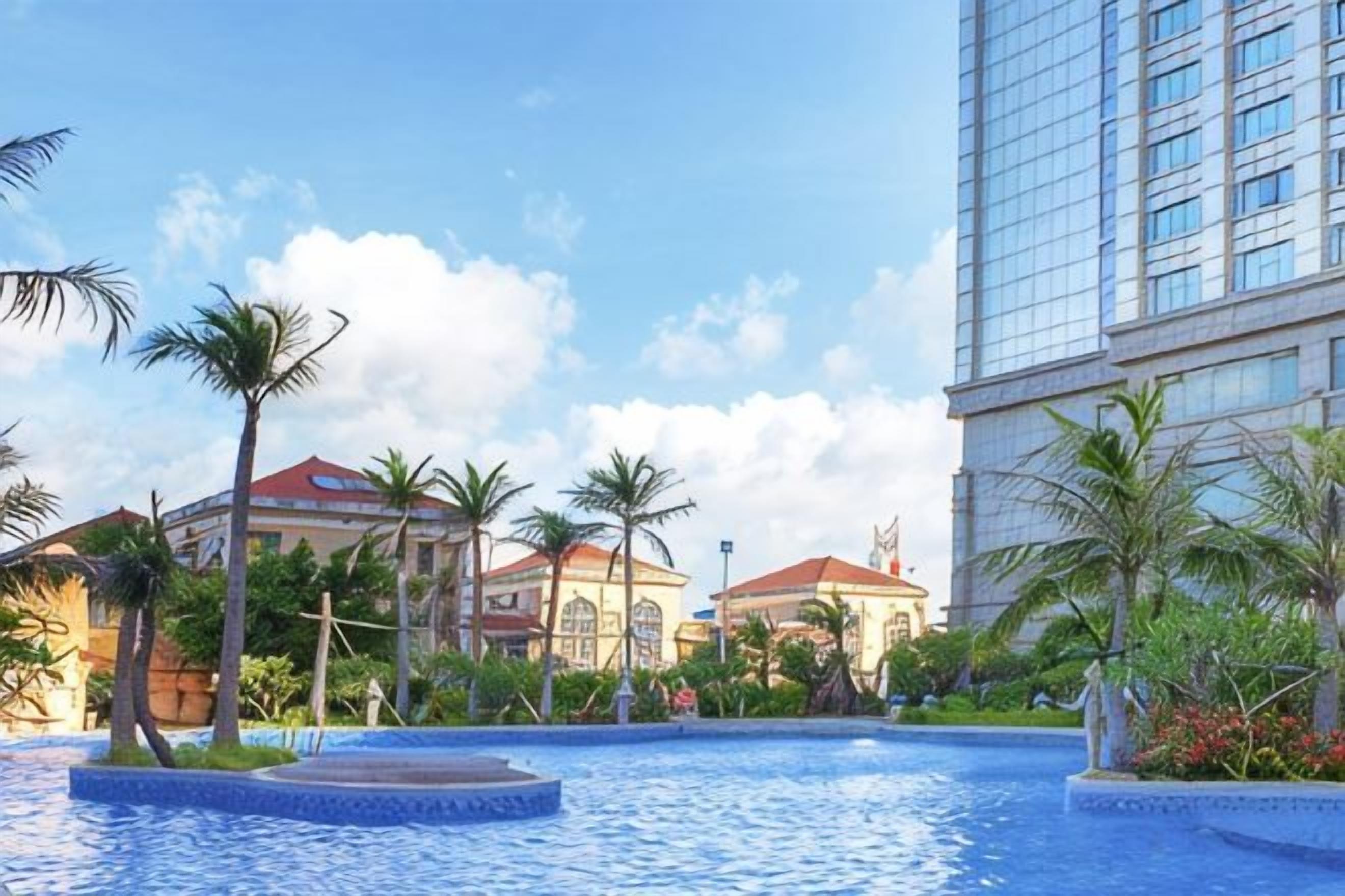 A swimming pool with a fountain, surrounded by palm trees and buildings with red roofs.