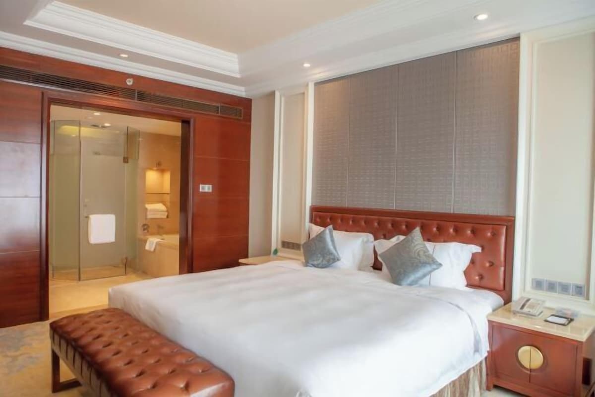 A hotel room with a large bed, a wooden bench, a nightstand, and a bathroom visible through an open door.