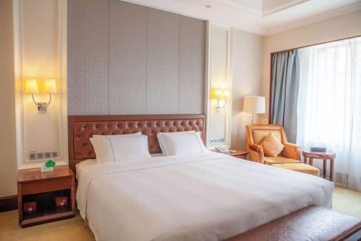 Quarto de hotel com uma cama grande, duas mesas de cabeceira, uma cadeira e uma janela com cortinas.