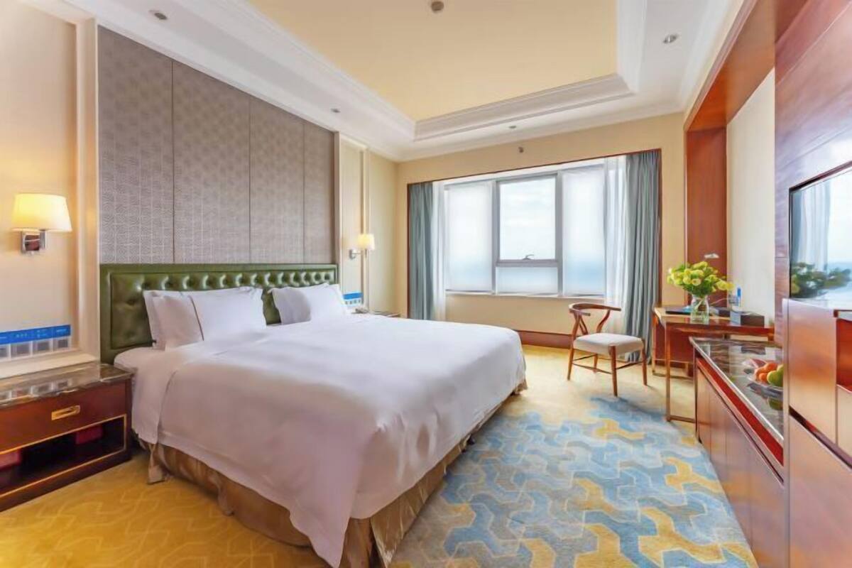 Quarto de hotel com uma cama grande, uma mesa com cadeira, uma janela com cortinas e vista para a cidade.