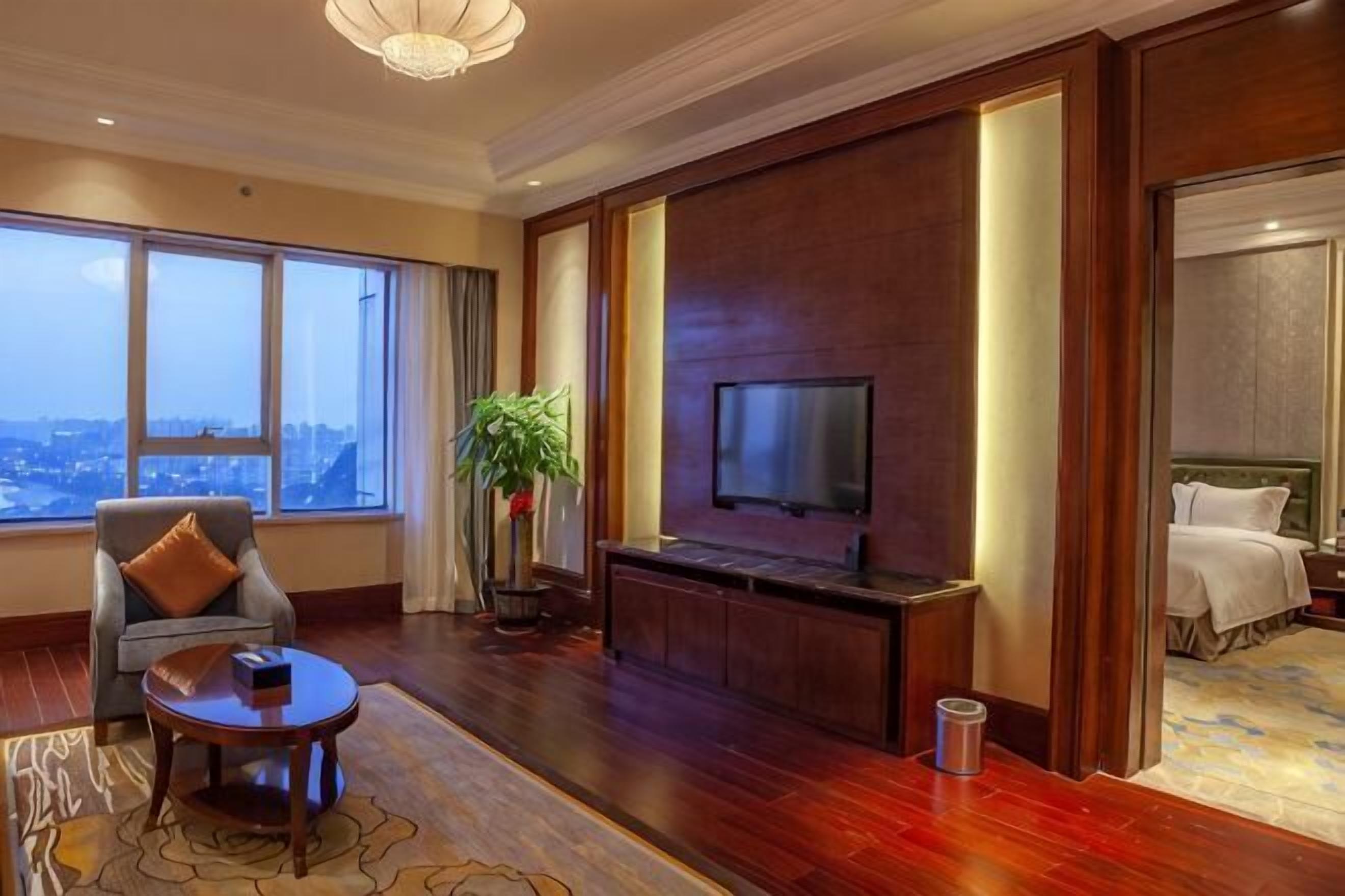 Habitación de hotel con televisor grande, zona de estar con una mesa, una planta y vistas a la ciudad.