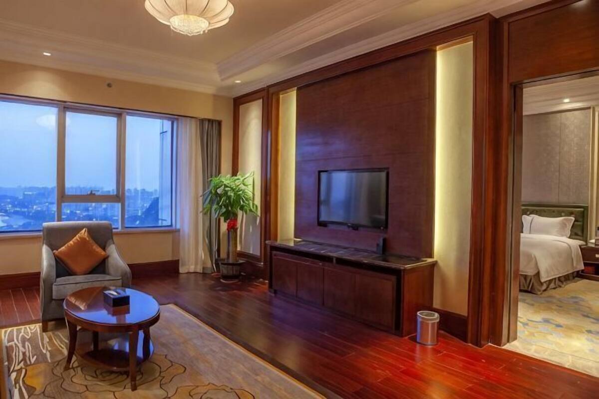 Quarto de hotel com uma televisão grande, área de estar com uma mesa, uma planta e vista da cidade.