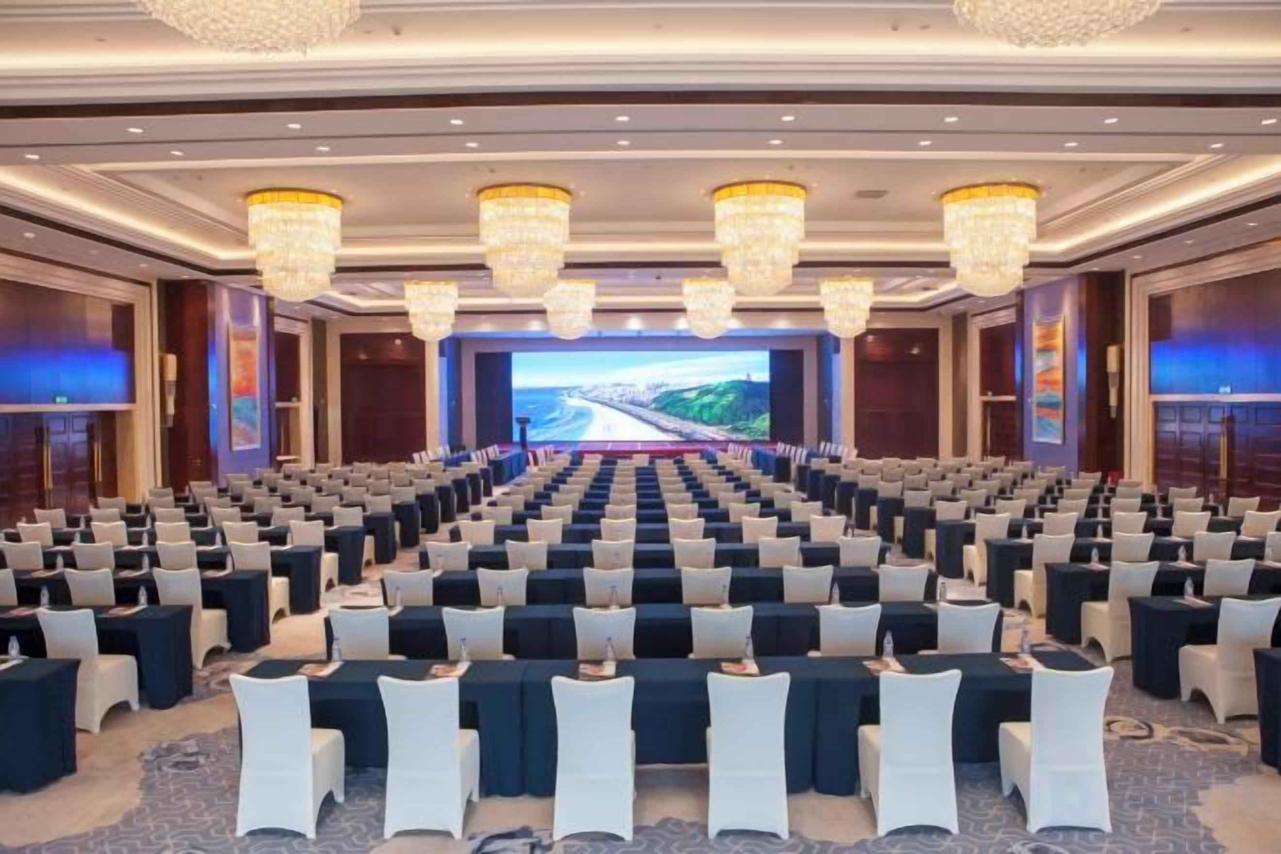 A conference room with rows of chairs, a large screen at the front, and chandeliers.