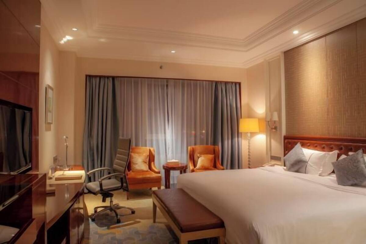 Quarto de hotel com uma cama grande, duas poltronas, uma escrivaninha com abajur, televisão e uma janela ampla com cortinas.