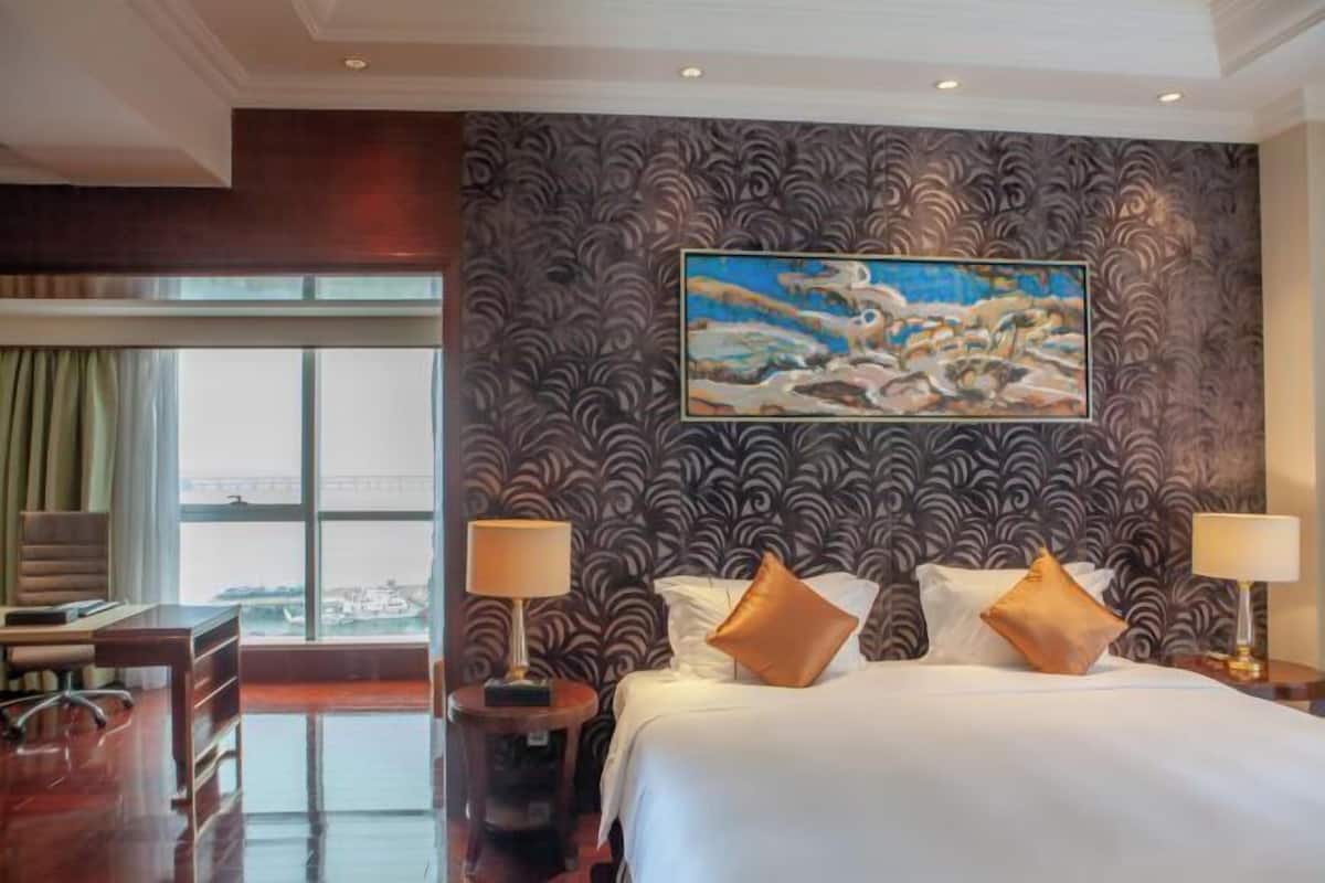 Quarto de hotel com uma cama grande, duas mesas de cabeceira com abajures, uma escrivaninha e uma cadeira. Há uma janela com cortinas e vista para a cidade.
