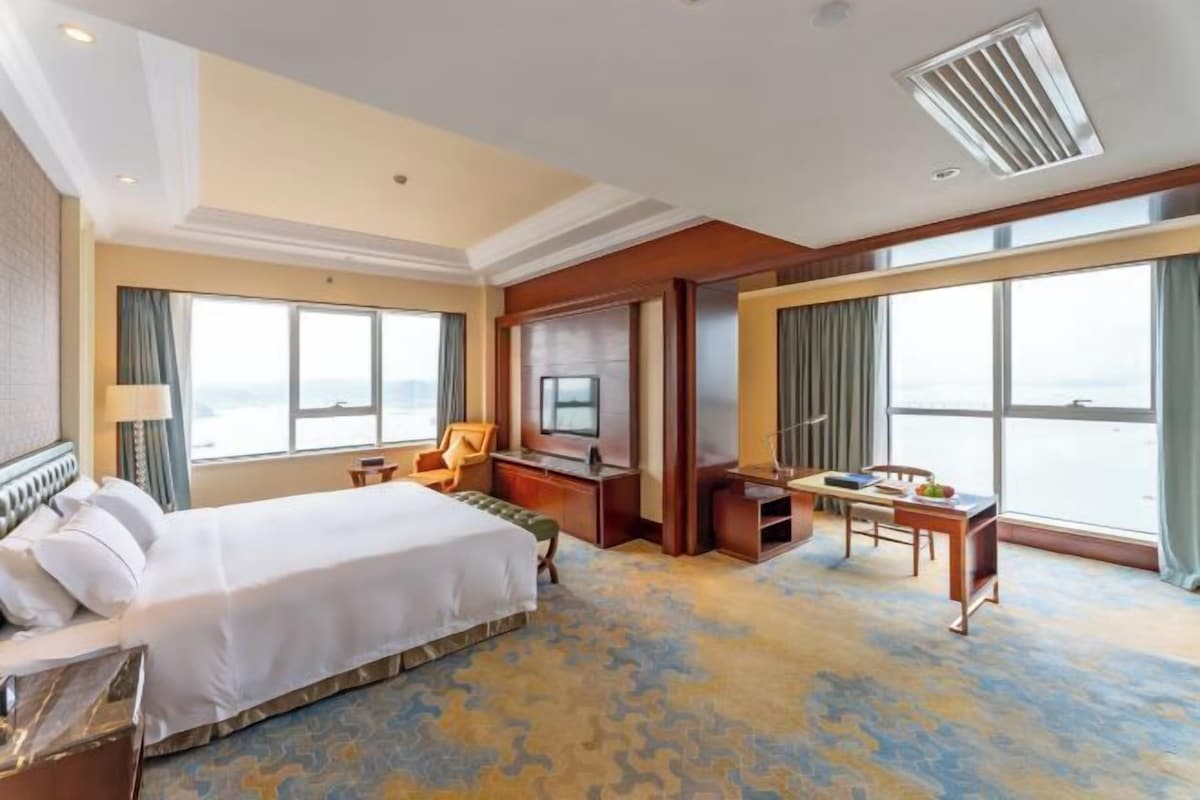 A hotel room with a large bed, a desk, a television, and a view of the sea.