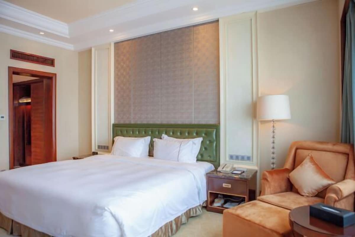 A hotel room with a large bed, a green upholstered headboard, a bedside table with a lamp, and a seating area with a chair and ottoman.