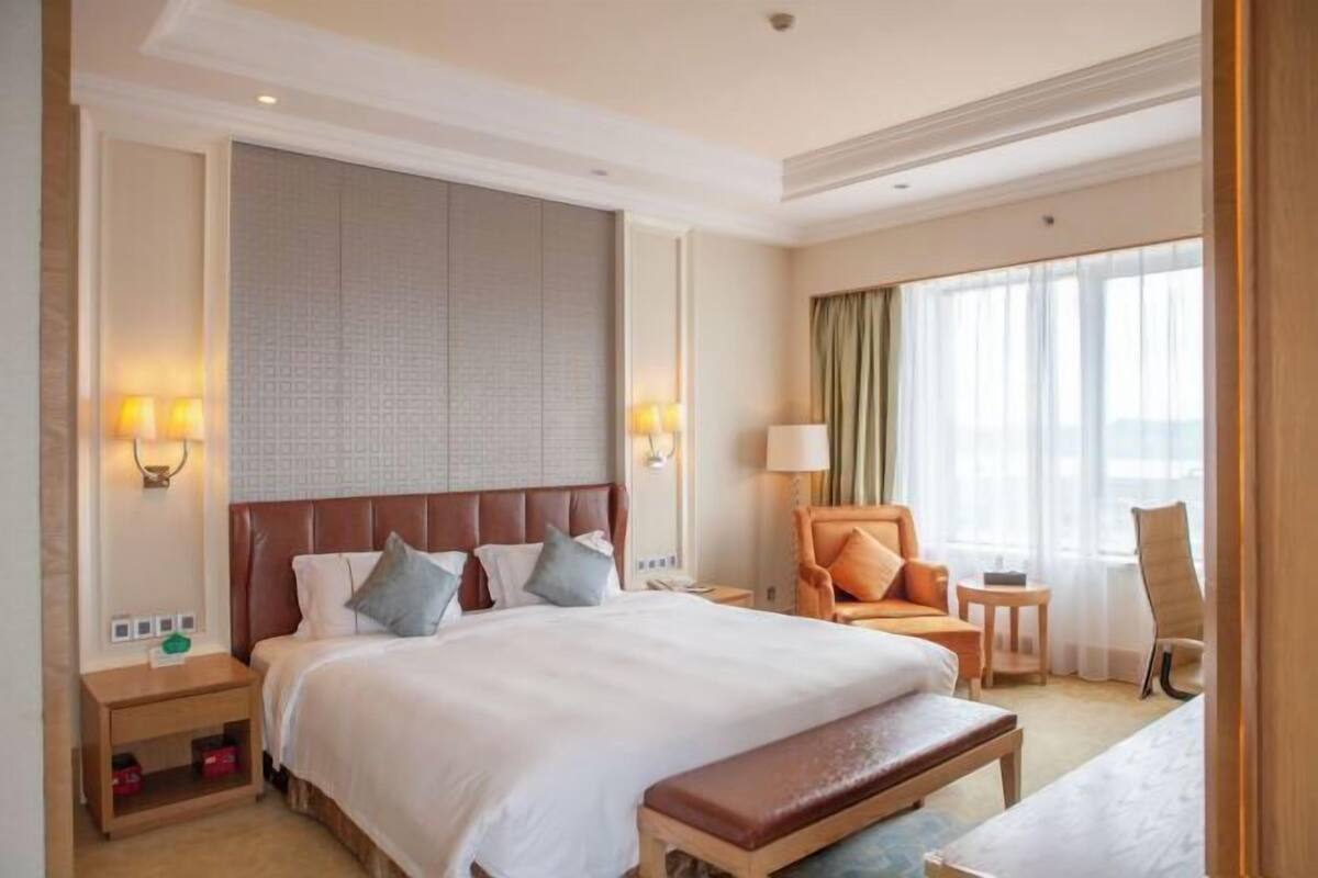 Quarto de hotel com uma cama grande, um banco, duas cadeiras, uma mesa de cabeceira e uma luminária fixada na parede.