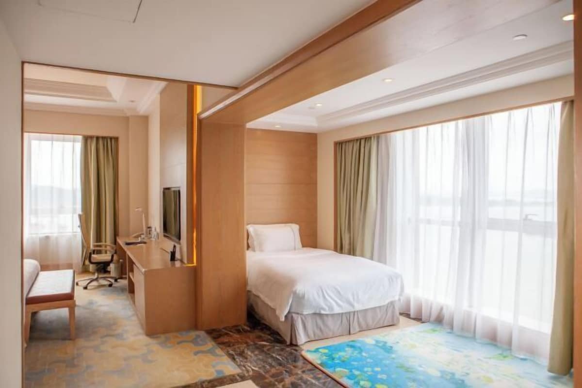 A hotel room with a bed, a desk, a chair, a television, and large windows with curtains.