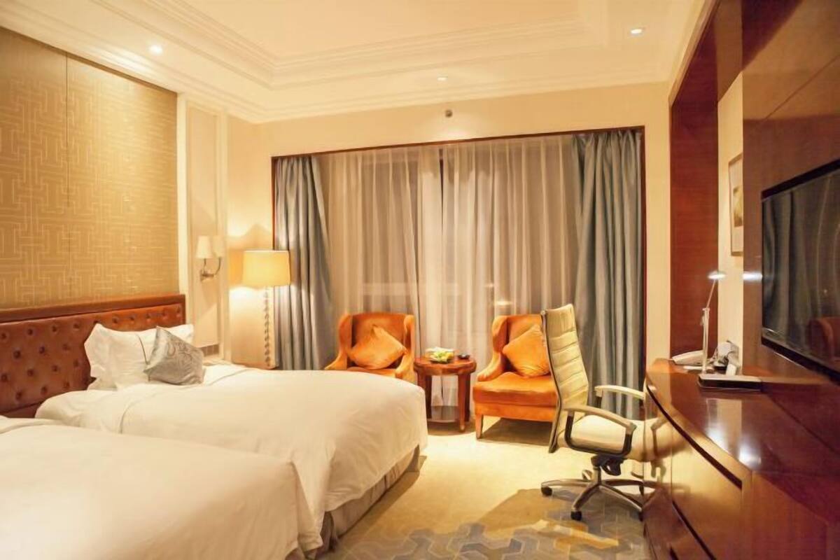 Quarto de hotel com duas camas, uma escrivaninha, uma cadeira, televisão e uma janela grande com cortinas.