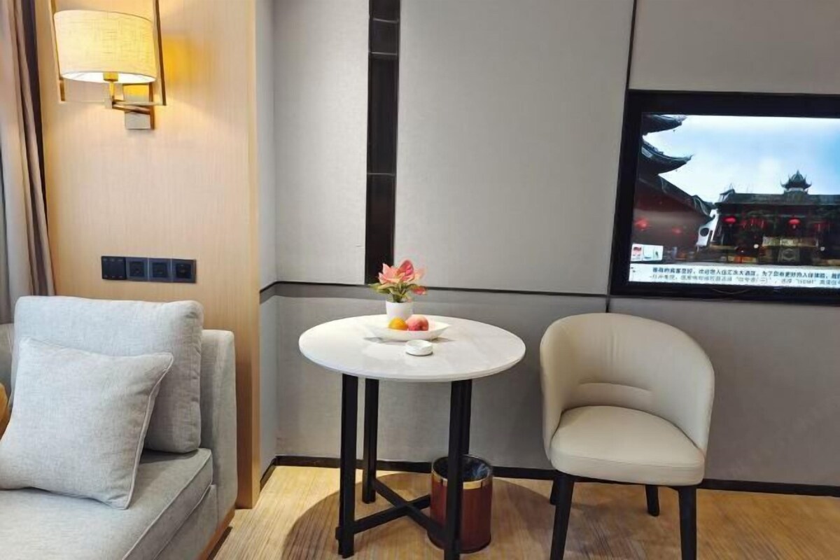 A hotel room with a sofa, a small round table with a flower vase, a chair, a TV mounted on the wall, and a trash bin.