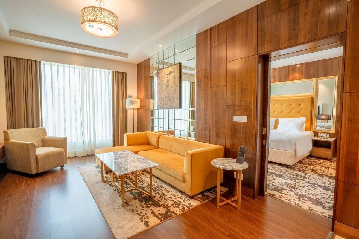 Habitación de hotel con sofá, sillón, dos mesitas, cama con cabecera, espejo y un ventanal con cortinas.
